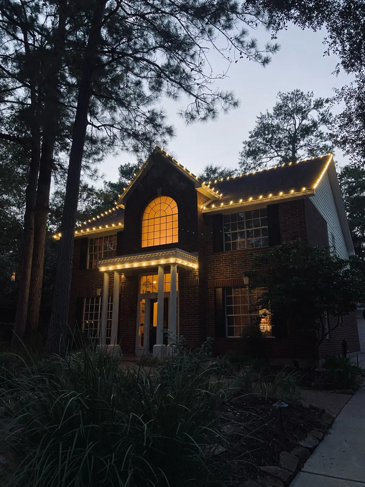 A large stucco estate at twilight with warm white C9 bulbs outlining every roofline and the arched front window, above a stamped-concrete circular drive