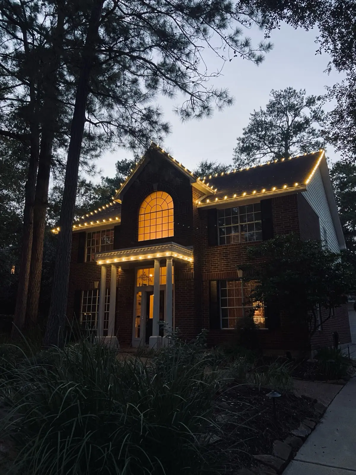 A two-story brick home at dusk with warm white C9 bulbs custom-cut along each gable and the front porch
